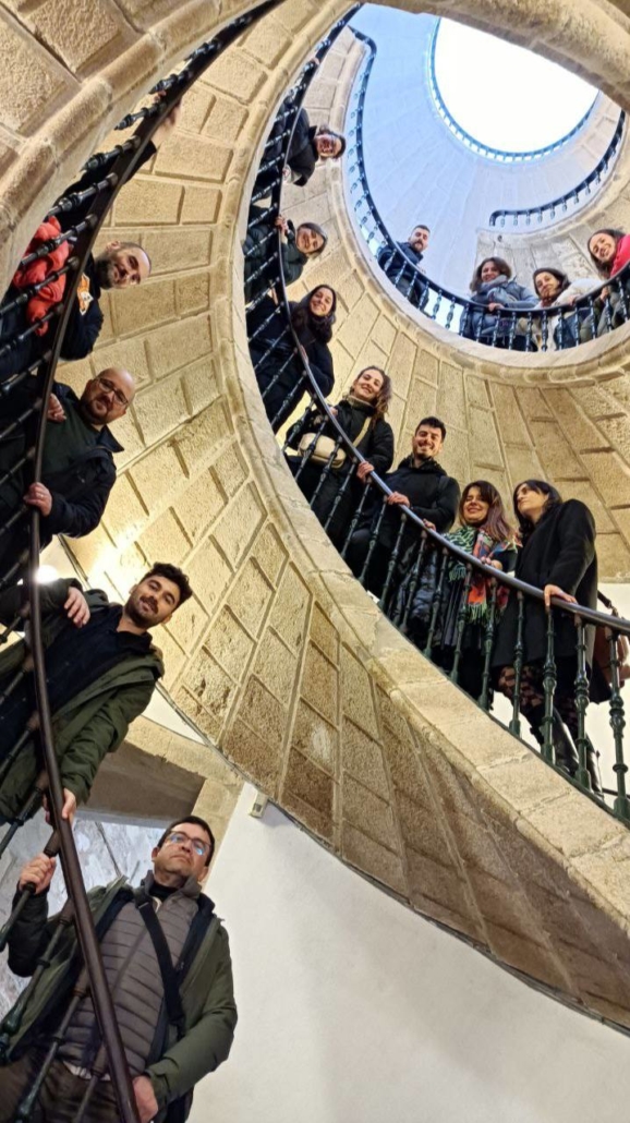 Foto vertical e contrapicada, dende a base da escaleira helicoidal do Museo do Pobo Galego, co alumnado da sétima edición do Curso repartido nos chanzos da escaleira